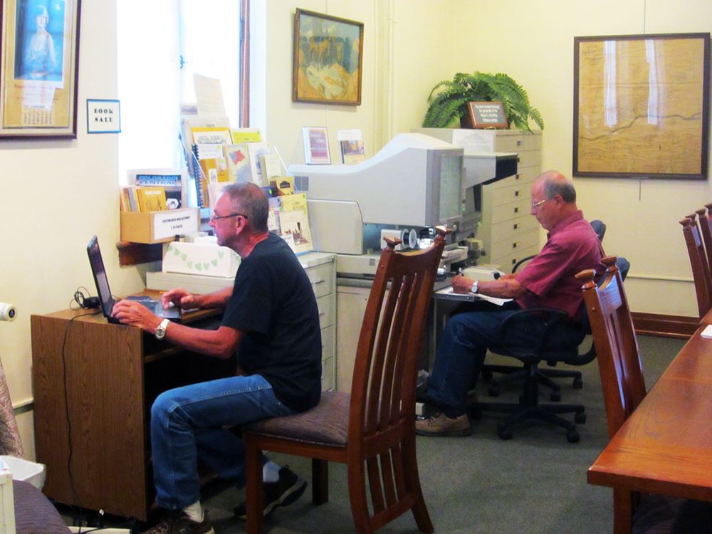 Color photo of two museum volunteers working at computer and mircro-film machine.