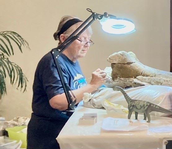 Color photo of museum volunteer Loretta Bailey working on a dinosaur fossil. Circa 2023.